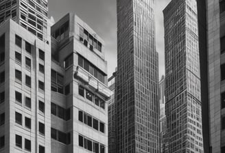 A modern, uniquely designed building with multiple reflective glass panels that create a checkered pattern. The upper parts display the names 'Norton Rose Fulbright' and another in Chinese characters. The structure exhibits a futuristic architectural style with sharp angles and a mix of straight and curved lines. In the foreground, additional buildings are visible with balconies and greenery, suggesting an urban setting.