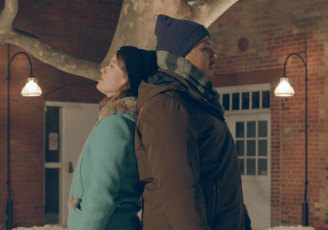 A young woman and man in winter coats stand with their backs together.