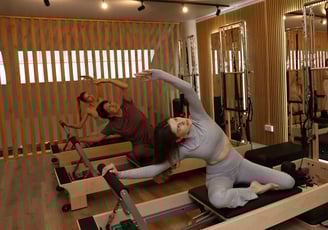 Três pessoas a fazer um alongamento lateral em reformers num estúdio de Pilates com espelhos 