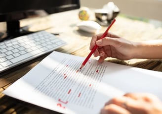 a person proofreading and correcting a document with a red pencil, computer in the background