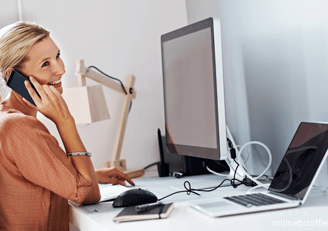 a woman talking on the phone phone while sitting at a desk with desktop and laptop