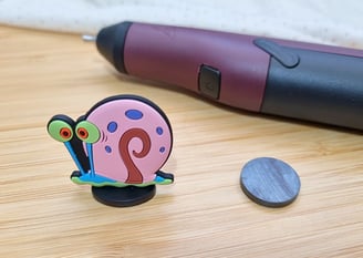 A colorful snail figurine with big eyes next to a hot glue gun and a round magnet on a wooden surfac