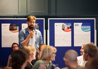 Ein blonder Mann mit Bart stellt bei einer Veranstaltung eine Frage.