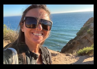 Michael Alexa Pitts on a rocky Pacific coastline with ocean behind, smiling, with black sunglasses & strands of hair in face