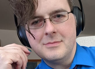 photo of GJ, white adult, with asymetrical wavy hair, glasses, chunky headphones, graying temples, nailpolish, & paisley tie
