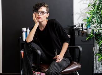 A hairdresser sits in a salon chair holding posing with a pair of scissors