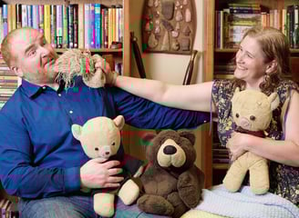 a man and woman sitting on a chair with teddy bears fighting each other