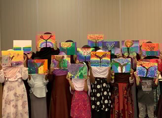 a group of Muslim girls standing in rows, holding colorful tree paintings up, hiding their faces