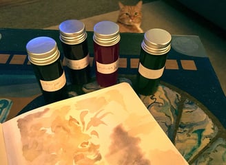 An open sketchbook with a fantasy watercolour illustration on a table surrounded by inks and a cat.