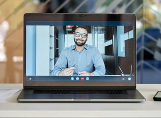 A laptop screen showing a video call with a man in a business shirt holding a pen