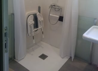 Une salle de bains pour personne à mobilité reduite, avec un siège ddouche rabattable, barres de maintient et une paroi à mii