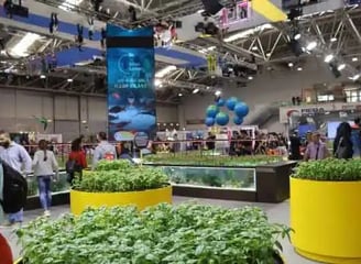 An indoor exhibition or trade show filled with people walking around various displays. Large planter