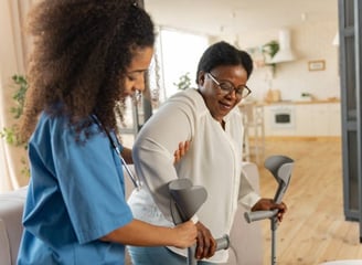 A woman using crutches stands beside another woman using a walker, both smiling in a supportive envi