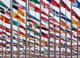 A border checkpoint features several lanes and a white structure with multiple signs indicating passport control. Vehicles, such as cars and a bus, are visible, and there are people both inside and outside the cars. The setting is outdoors under a cloudy sky, and European Union flags are seen above the building.