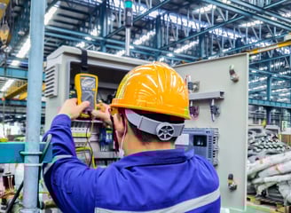 Industrial electrician wearing hard hat and gloves, servicing a large electrical panel in a warehous