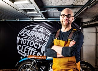 Homme barbu et avec des lunettes debout devant une moto souriant