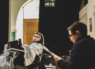 An electric wheelchair user with ventilator mask and a white BSL signer deep in conversation