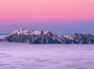 a snow covered mountain range poking above the clouds shrouded in pink purple evening sky