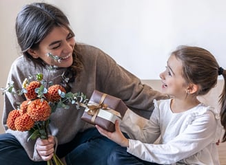 Niña con camisa blanca dando regalo a mamá con sueter gris y sostentiendo flores