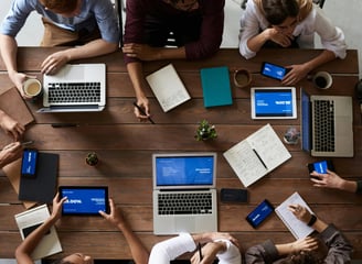A group of people sitting around a table with laptops, pens, notebook, and cups.