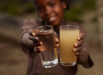 A smiling young girl holding two bottles, showing the difference between dirty and purified drinking