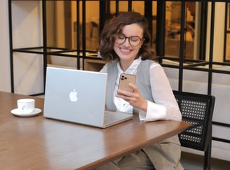 35+ woman mentor smiling, wearing gray, holding phone, sitting with MacBook on table