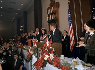 JFK in a suit and tie standing at a podium with a microphone
