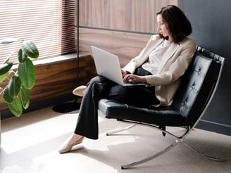 A business woman in a cream jacket and dark slacks,sitting on a black chair, working on a laptop.