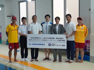 イトマンスイミングスクールと日本ライフセービング協会が『水辺の事故ゼロ』に向けたパートナーシップ協定を締結し、関係者が協定書を持って並んでいる様子