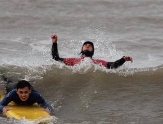 A prone smiling surfer dropping down a wave while a surf instructor jumps backwards over a wave.