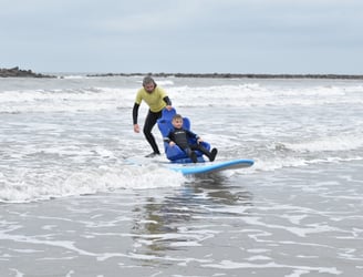 Young surfer in a seated surfboard with a coach controlling the board riding a wave