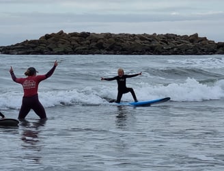 A young male surfer riding a wave cheerring as a female surf coach gives him encouragment.