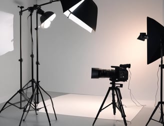 A well-lit film or photography studio setting with several people working. The backdrop is an expansive white screen with lighting equipment overhead and on the sides. Individuals are scattered throughout, some appearing to adjust equipment or collaborate on tasks.