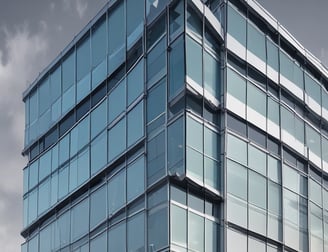 A modern office building with a prominent glass facade and a large colorful logo featuring a letter 'T' on the upper section. The entrance is covered by a transparent canopy supported by metal beams, leading into the building. The architecture displays a sleek, contemporary design with a mix of glass and concrete elements. Large windows are visible on both sides of the structure.
