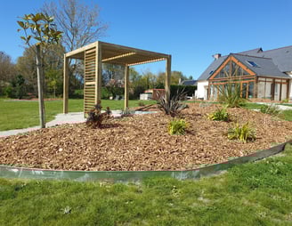 Massif paillé avec jeunes plantations devant une maison bois et pergola en fond