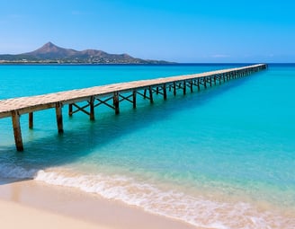 Playa de Muro en Mallorca con muelle de madera sobre aguas turquesas y montañas al fondo – Maiorca Unlocked.