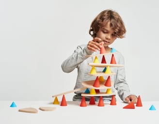 A boy creating a structure from red, blue and yellow silicone cones and wooden tables in the room