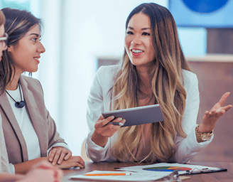 Asian business consultant discusses data while holding a tablet in front of three other female leaders in a boardroom.