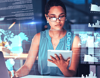 A female leader with glasses holds a tablet and types on a laptop at her desk, surrounded by holographic maps and graphs.