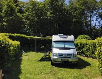 Spacious campervan pitch on grass with awning and outdoor table, surrounded by hedges and forest