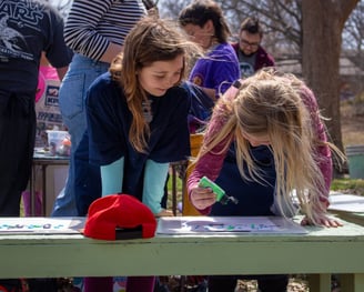 Two young girls smiling while standing over papers dripping paint onto them.