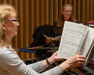 a woman in glasses and a gray sweater playing a piano