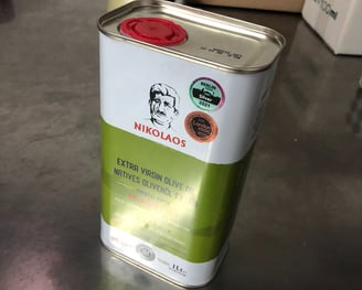 Eine Dose NIKOLAOS Extra Virgin Olive Oil steht auf einer metallischen Oberfläche, mit Preissiegeln 