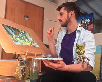 A handsome young male artist with dark hair and blue shirt sits at his easel of fantasy art