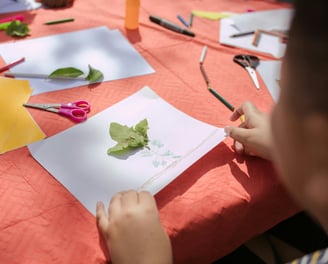 a child's hands are holding scissors and paper and leaves are on the table