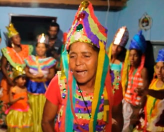 Mulheres e crianças com trajes e coroas coloridas cantam e dançam o Reizado em uma casa decorada