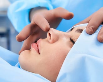 Patient draped in blue, surgeon's hands examine nose for rhinoplasty.