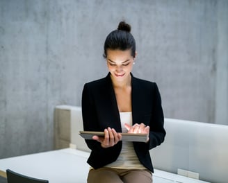 A businesswoman in a black jacket, sitting on a desk, holding a tablet computer.