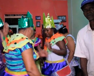 Grupo de mulheres em trajes do Reizado dançando com coroas coloridas ao lado de um homem observando
