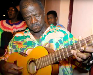 Sr. João Reis toca violão durante o Reizado, vestindo camisa florida, símbolo da tradição quilombola